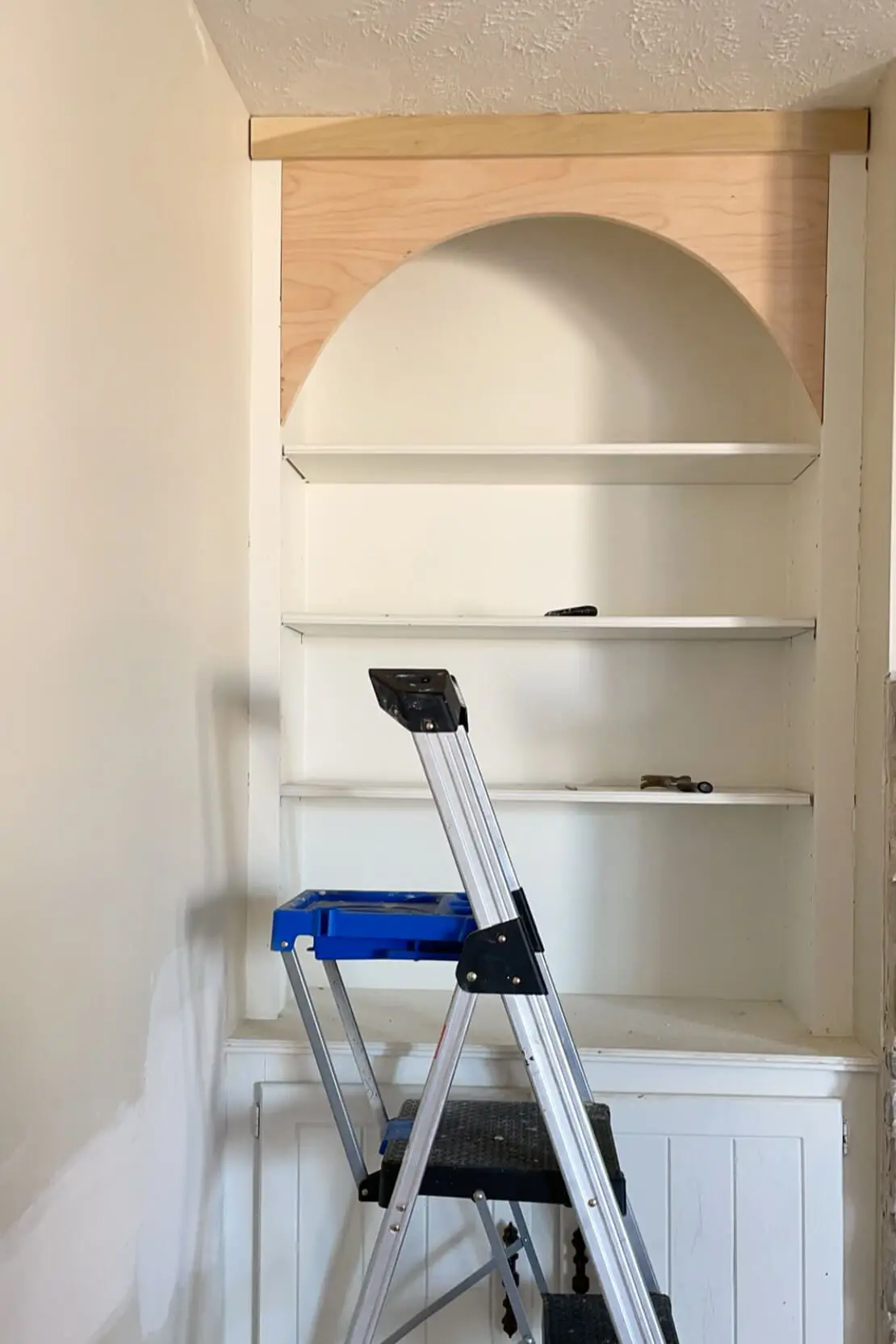 A built-in shelving unit in progress, featuring an arched detail added to the top with unfinished wood, ready for painting or staining. A blue step ladder is positioned in front, showing active DIY work on the project.